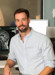 Timothy Hughes seated indoors at a workstation, wearing a light blue button‑down shirt, with medical imaging displayed on computer monitors in the background.