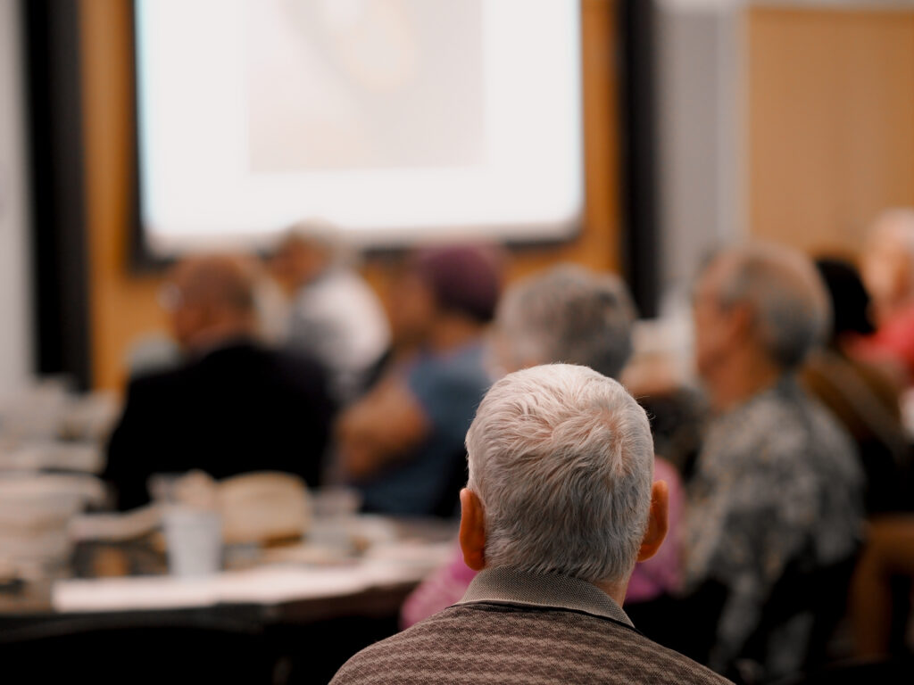Attendees sit at tables and watch a presentation during a community education event.